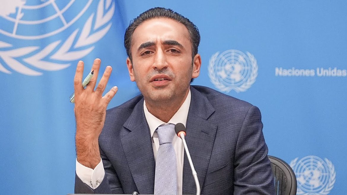 A man in a blue suit, with a light blue tie, speaking at a press conference in front of the United Nations backdrop, gesturing with his left hand while holding a pen in his right hand.