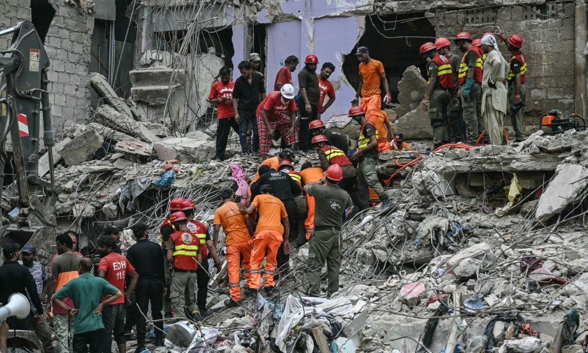 051623499739af8 The image depicts a rescue operation at the site of a collapsed building. Multiple rescue workers in orange and red uniforms are seen working amidst the debris, helping victims and searching for survivors. The scene shows the destruction of the structure, with exposed steel rods and rubble scattered around the area. Emergency responders are actively involved in recovery efforts, while some people from the community are also present at the scene.