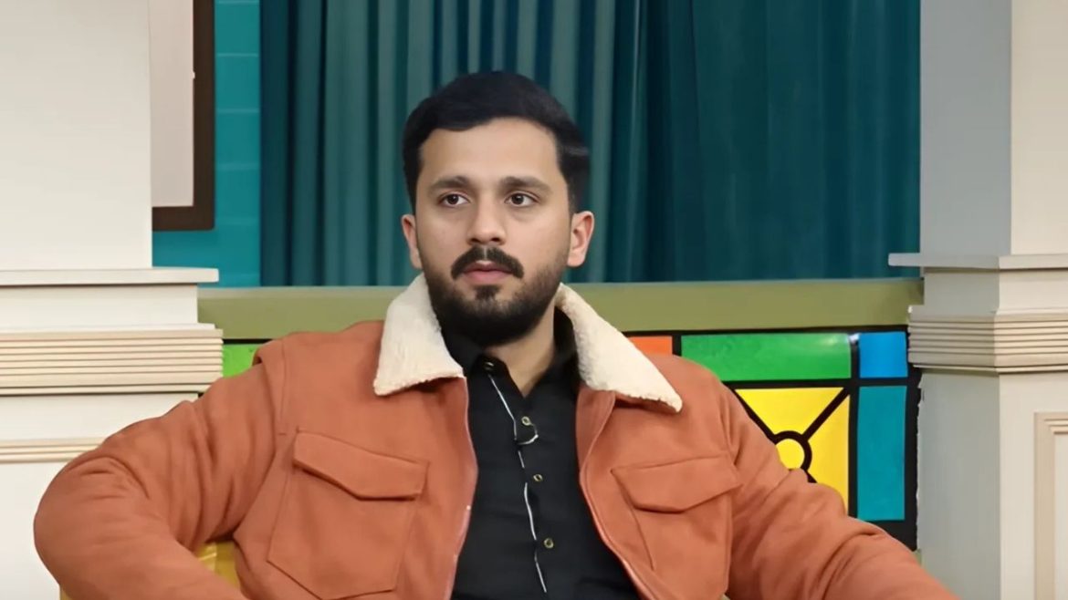 A man with a beard seated in a chair indoors, wearing outerwear.