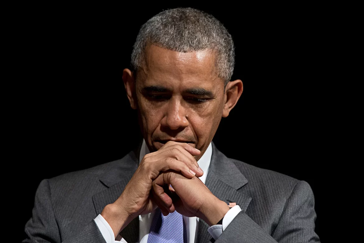Former U.S. President Barack Obama in a gray suit sits with hands clasped in front, against a dark backdrop, exuding a contemplative demeanor.