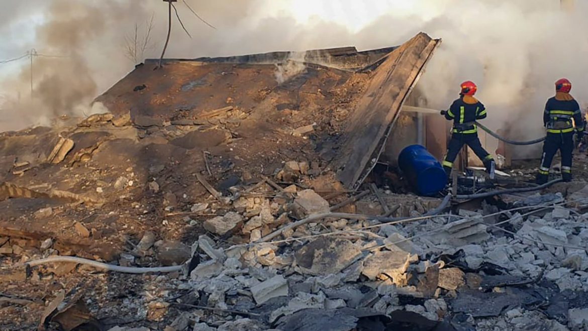 Two firefighters extinguish smoke from a collapsed building amidst debris, with cables overhead and a blue barrel visible.