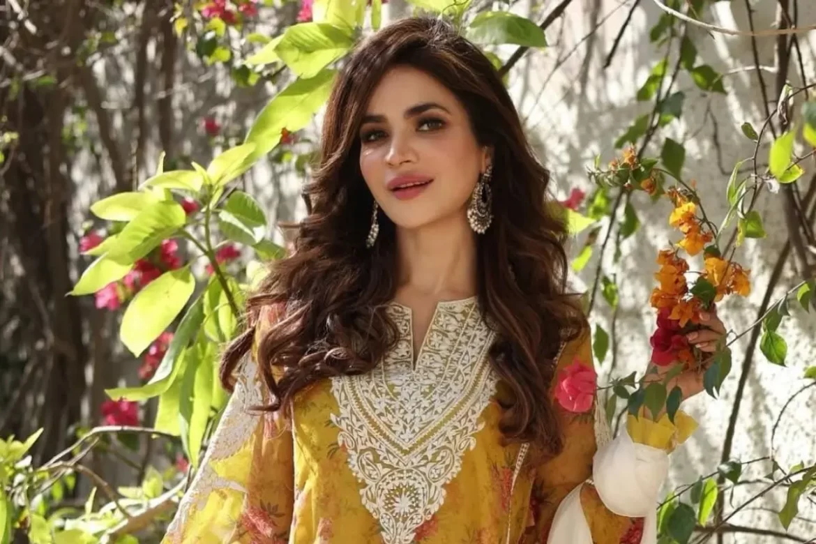 humaira-asghar-2-jpg A woman in a yellow embroidered outfit, with long brown hair, standing in front of a vibrant green and pink floral background. She is holding a branch with orange and pink flowers.