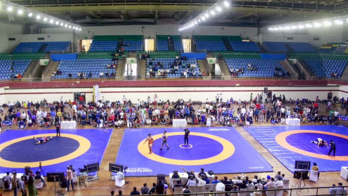 A spacious wrestling arena with blue and orange mats, featuring matches in progress, surrounded by seated spectators and coaches.