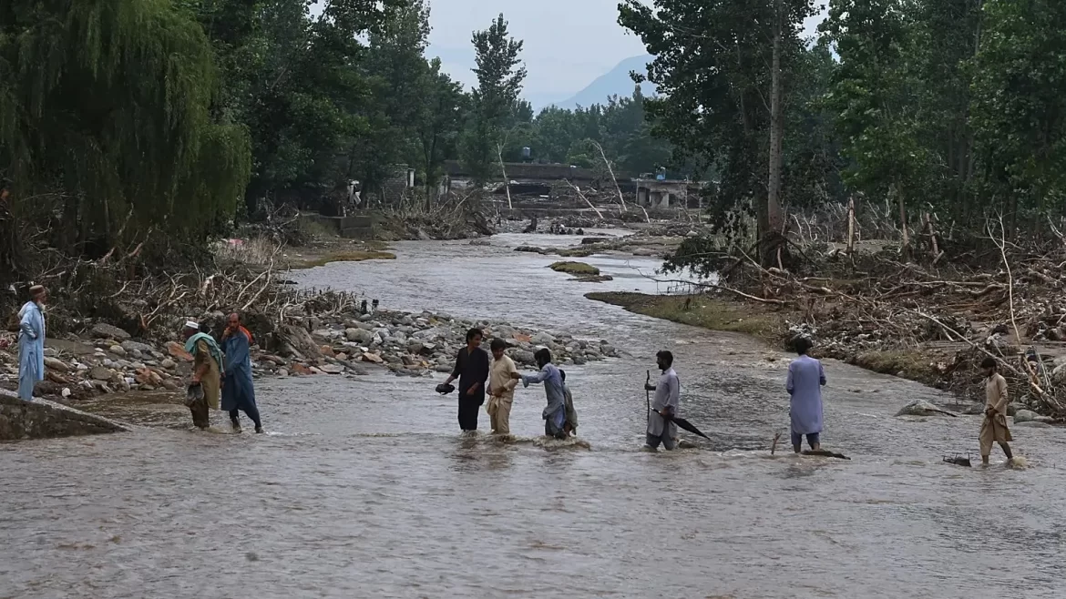 People navigating a flooded river, surrounded by debris and damaged trees, highlighting the aftermath of a natural disaster.