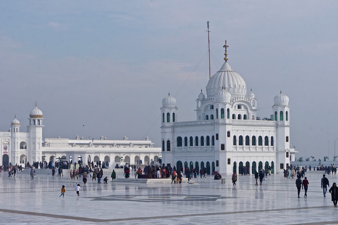 A large crowd gathers outside a stunning white Sikh gurdwara with domes and arches, set against a clear sky.