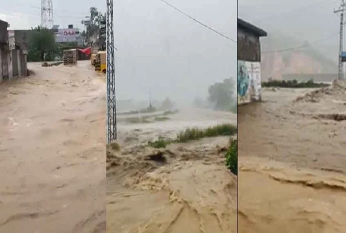 A flooded street with rapidly flowing muddy water, obscured visibility, and nearby structures amidst heavy rain.