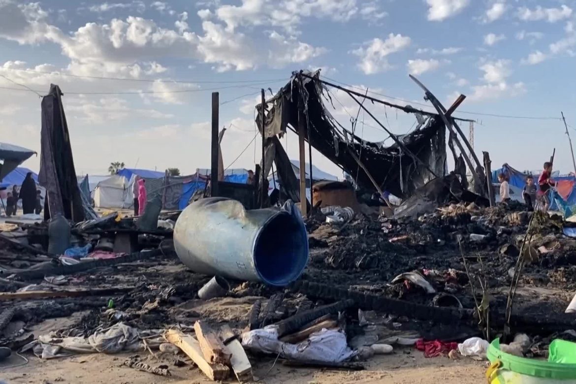 Devastated makeshift camp scene with burnt structures, debris, and survivors amid a smoky, damaged landscape under a cloudy sky.