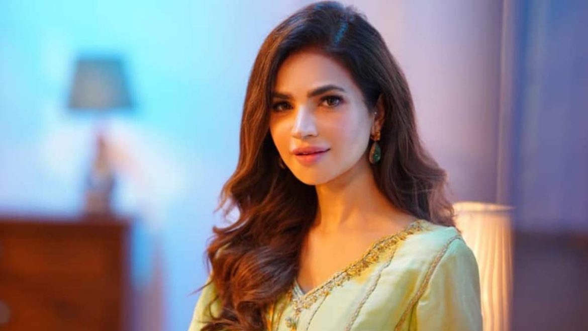 A woman in a light yellow embroidered outfit poses elegantly, with soft waves of brown hair against a softly lit blue background.