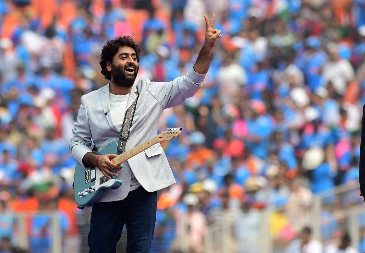 A musician in a light blazer raises two fingers while holding a guitar, with a vibrant crowd in blue attire cheering in the background.