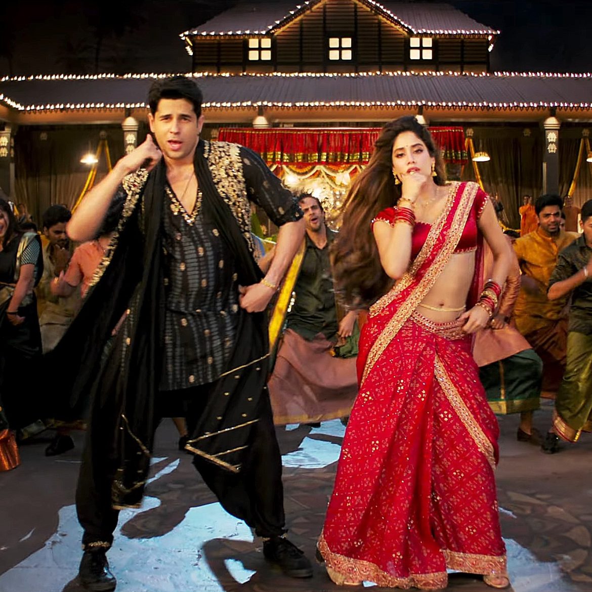A vibrant dance scene featuring a man in traditional attire and a woman in a red lehenga, surrounded by a festive group and decorated backdrop.