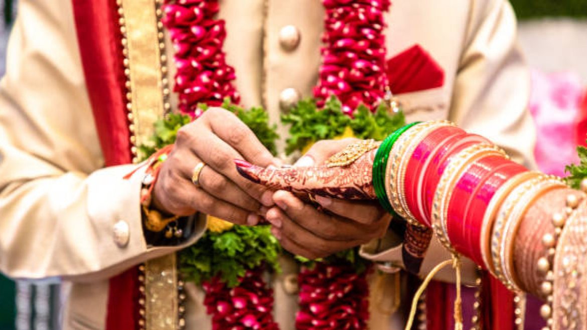 A groom in traditional attire holds a decorative wedding item, adorned with vibrant bangles and floral garlands, expressing cultural celebration.
