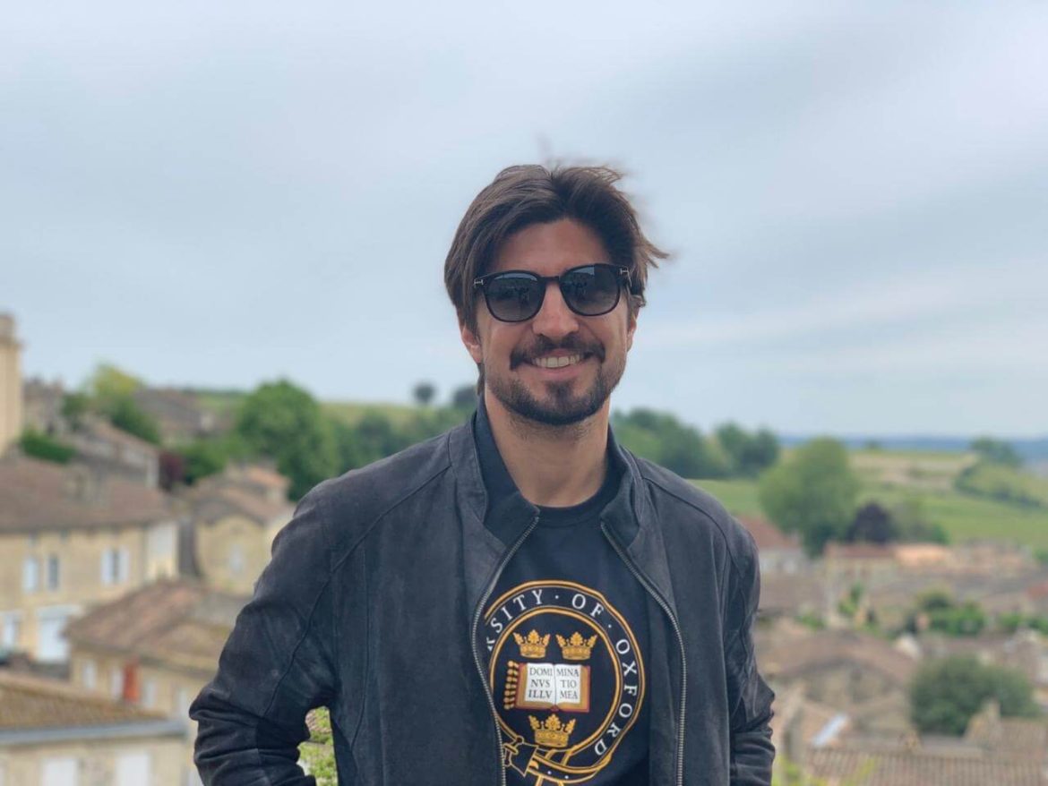 A person wearing a University of Oxford t-shirt stands against a scenic backdrop of green hills and buildings under a cloudy sky.