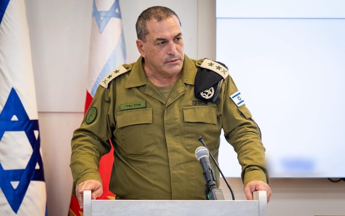 A military officer in a green uniform stands at a podium with a microphone, flanked by Israeli flags and a screen in the background.