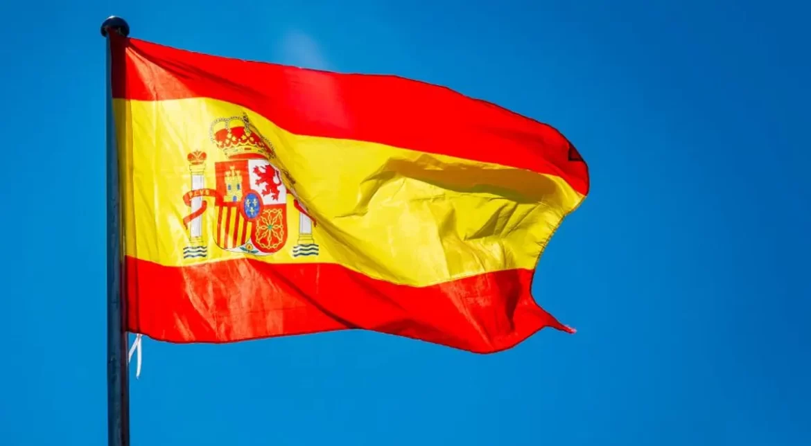 Spanish flag waving against a clear blue sky, featuring red and yellow horizontal stripes with the national emblem on the left.