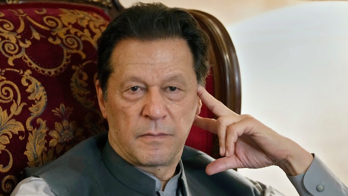 Former Prime Minister Imran Khan sits thoughtfully in an ornate chair, dressed in traditional attire, with a hand resting against their face.