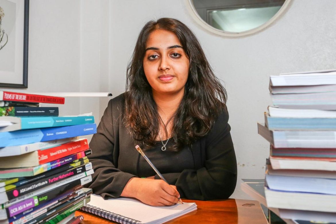 A girl sits at a desk surrounded by stacks of books, writing in a notebook with a pencil. Natural light illuminates the workspace.