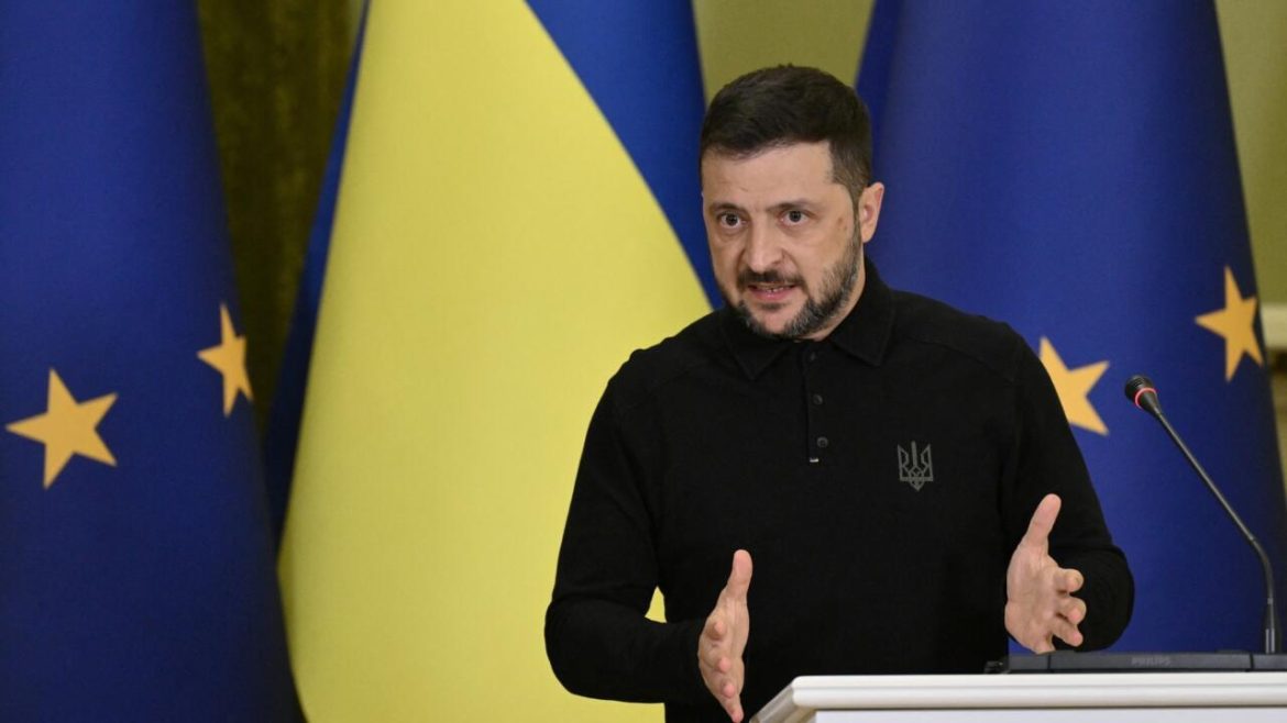 A person in a black polo shirt gestures while speaking at a podium, with blue and yellow flags in the background.