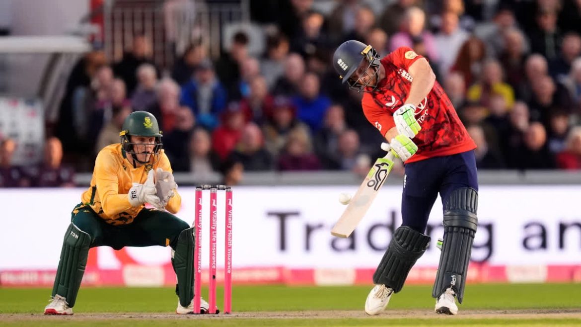 A cricket player in a red jersey swings a bat as a wicketkeeper in yellow crouches behind pink stumps, with a crowd in the background.