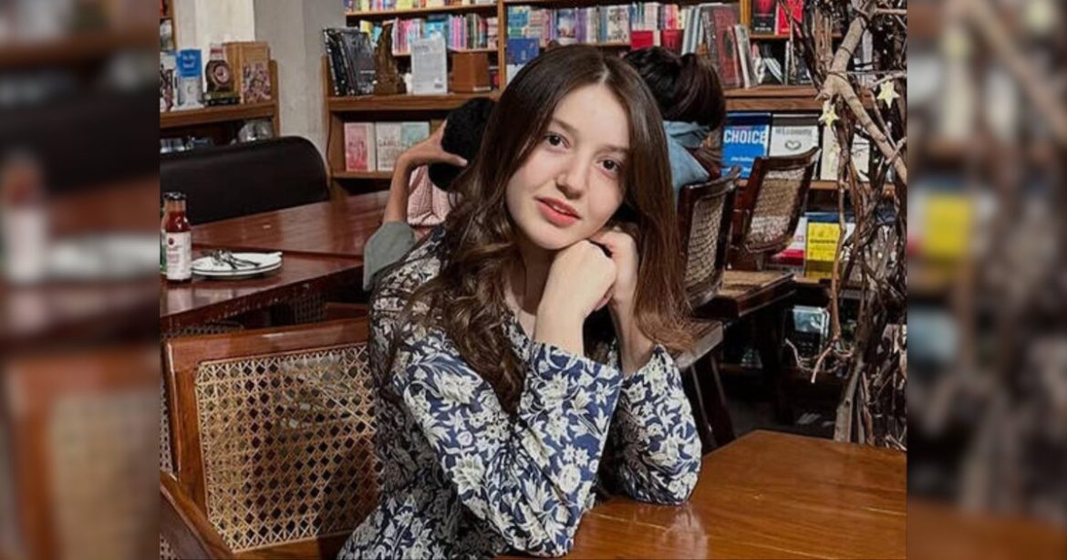 A woman with long, wavy hair sits at a wooden table in a cozy café filled with books, resting her chin on her hand, looking contemplative.