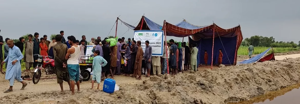 A crowd gathers near a makeshift tent with a banner, seeking assistance amidst flood-affected landscapes and cloudy skies.