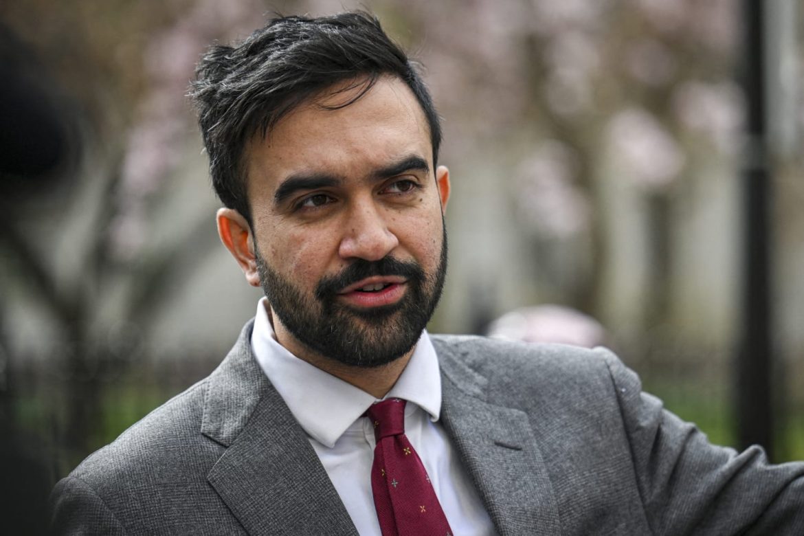 A man in a gray suit and a red tie gestures while speaking outdoors, surrounded by blooming trees and a blurred background.