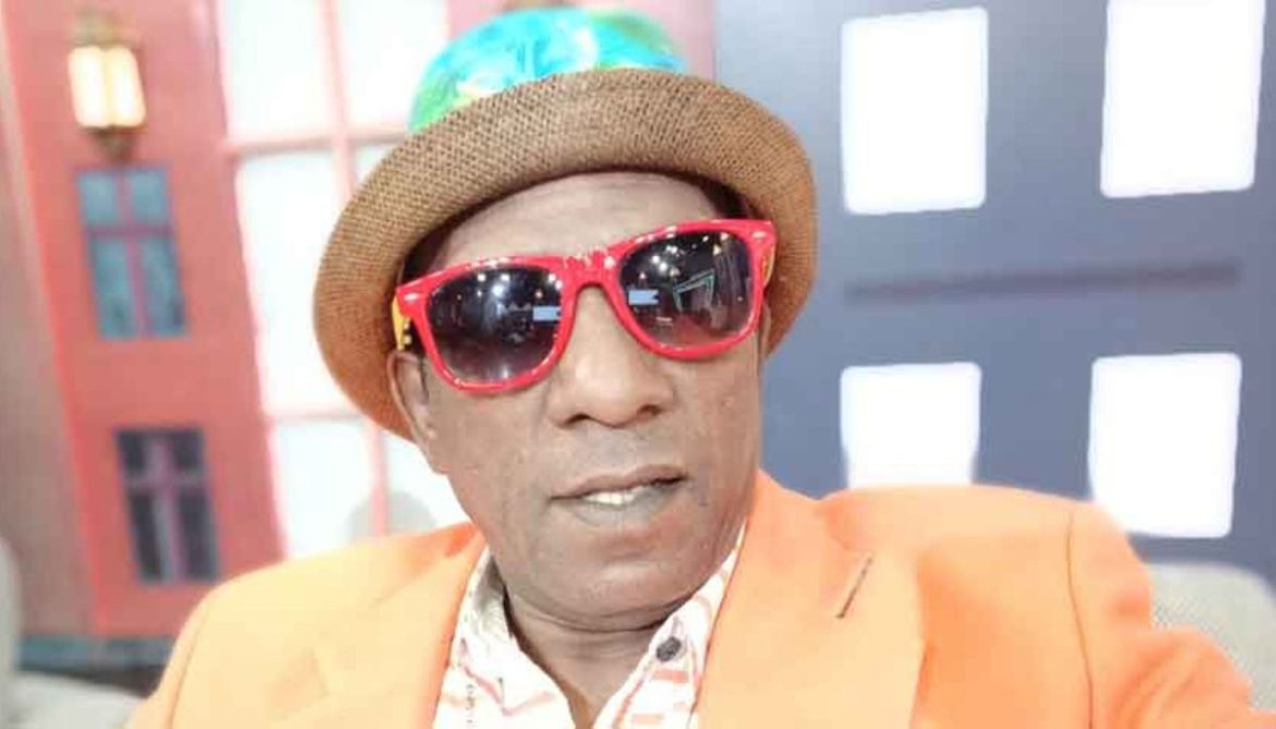 A person wears a vibrant orange blazer and a straw hat with colorful accents, seated in a cozy space with a colorful backdrop.