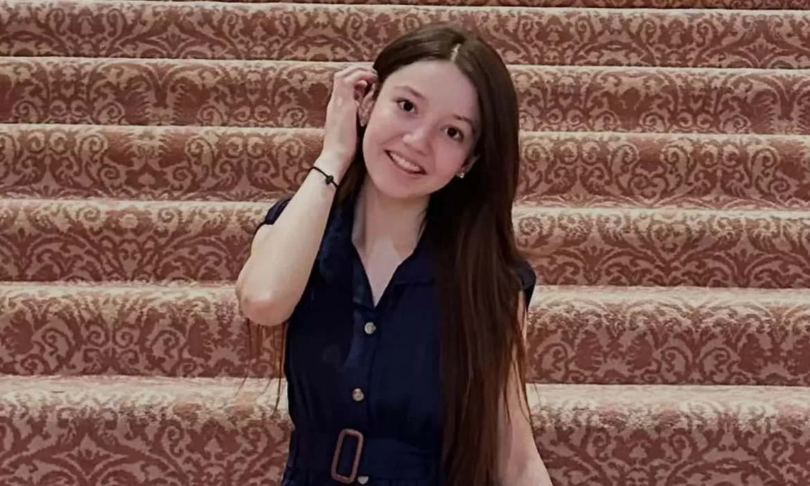 A person with long hair in a dark dress poses on a patterned staircase, holding their hand to their face.