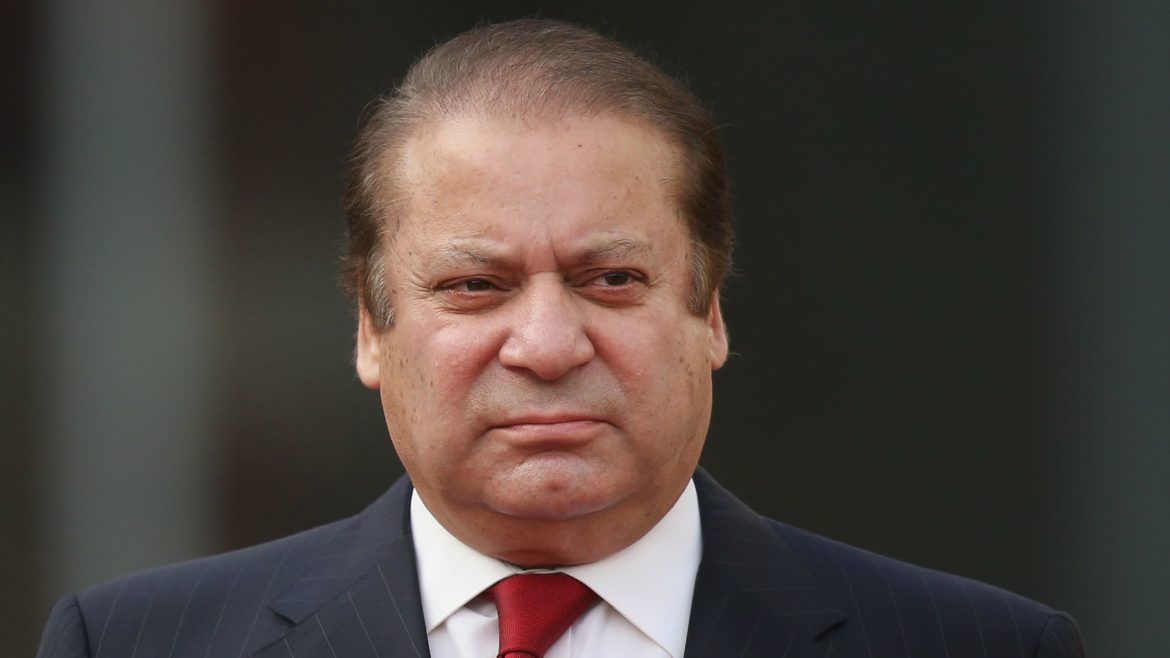 A middle-aged man in a formal dark suit and red tie stands against a blurred background, exuding confidence and authority.