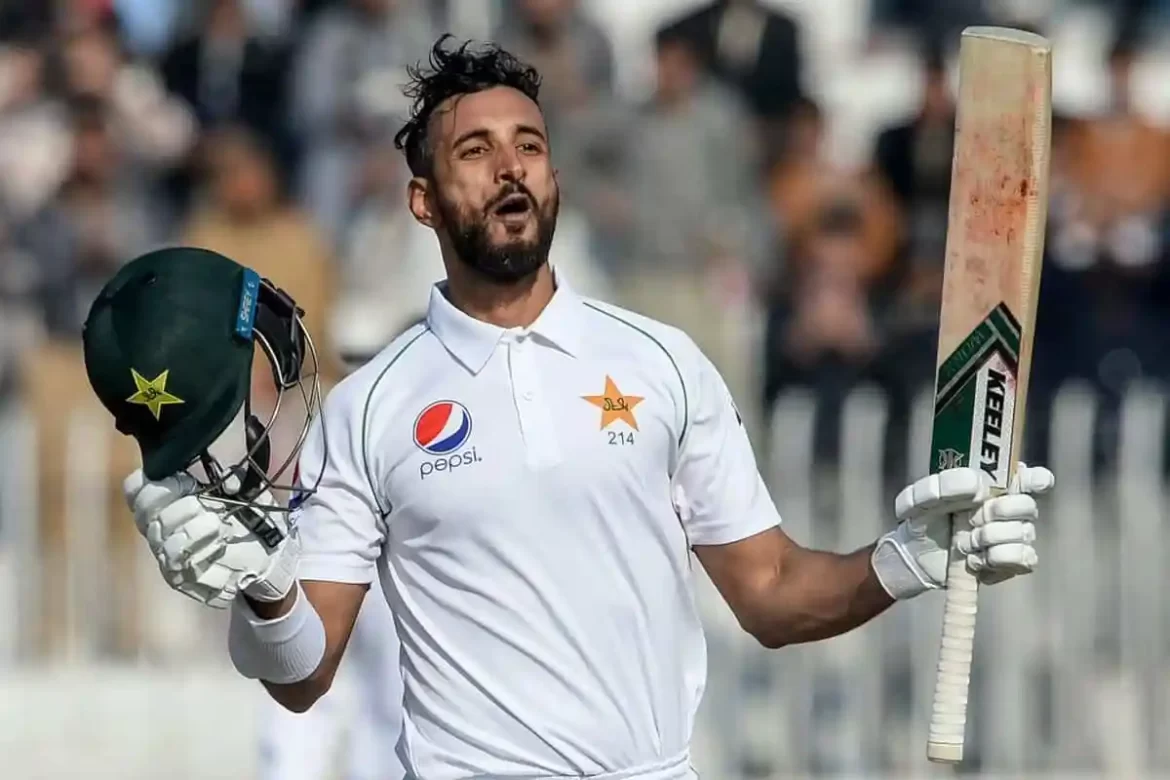 A Pakistani cricketer, in a white kit, holds a bat and helmet, celebrating a significant achievement on the field.