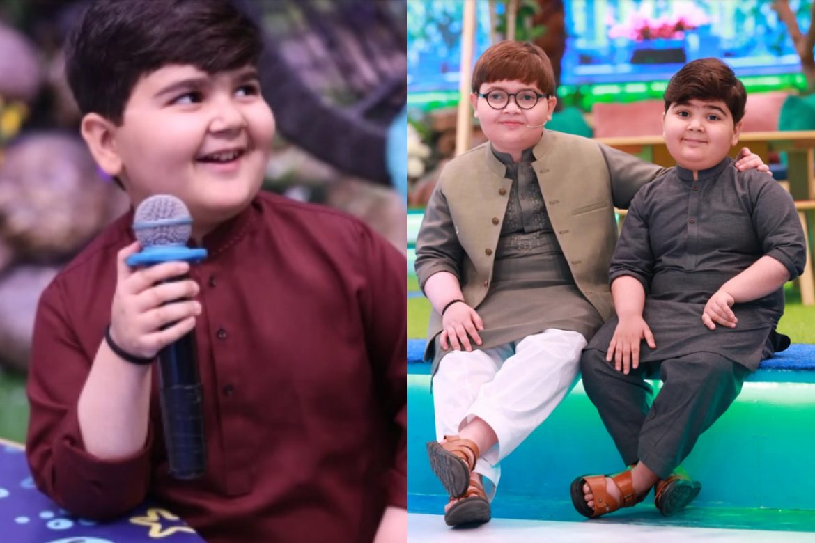 Two children sit together: one in a maroon shirt holding a microphone, the other in a traditional outfit with sandals, smiling.