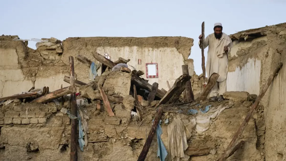 A person stands on the ruins of a collapsed structure, surrounded by debris and wooden beams under a clear blue sky.