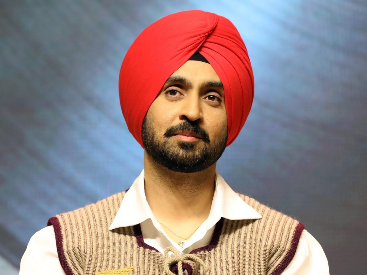 A person wearing a vibrant red turban and a beige sweater with maroon accents, set against a softly blurred background.