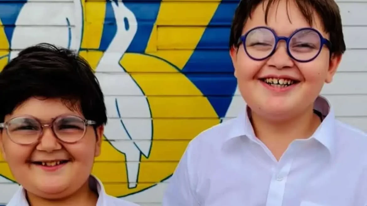 Two smiling boys wearing glasses, standing together in front of a colorful mural featuring storks. The boys are dressed in white shirts, with one of them having shorter hair and the other with longer hair.