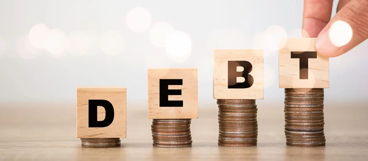 A hand places a wooden block labeled 'T' on stacked coins, spelling "DEBT" with other wooden blocks. Soft bokeh in the background.