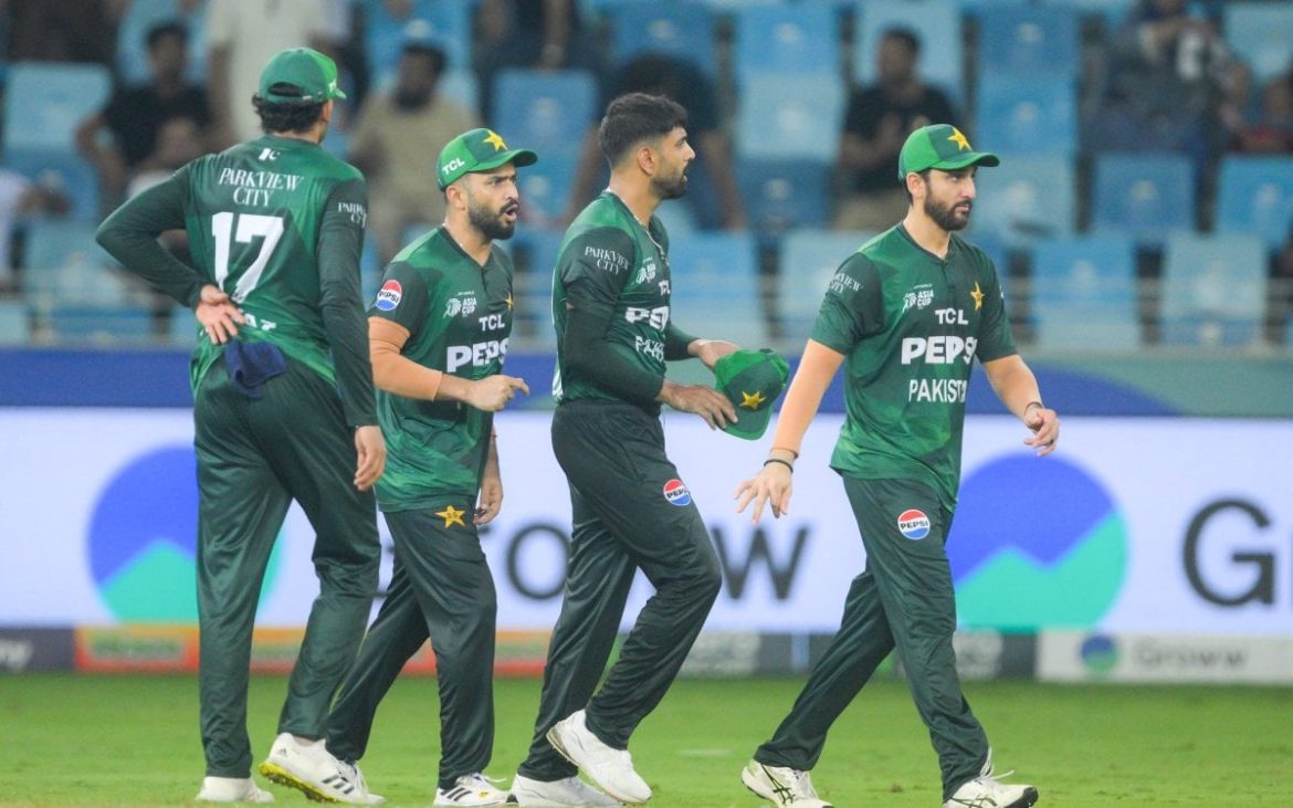 Four cricket players in green uniforms walk on the field, with a stadium backdrop and a crowd visible in the stands.