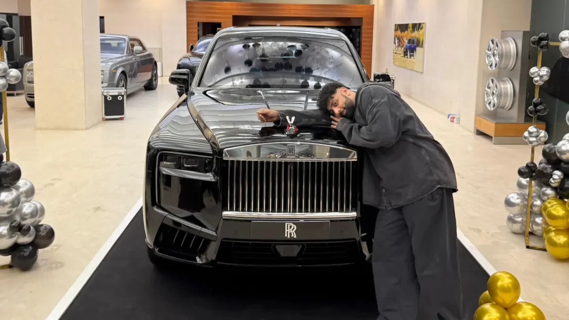 A person leans against a luxurious black Rolls-Royce in an elegant showroom, with decorative balloons and cars in the background.