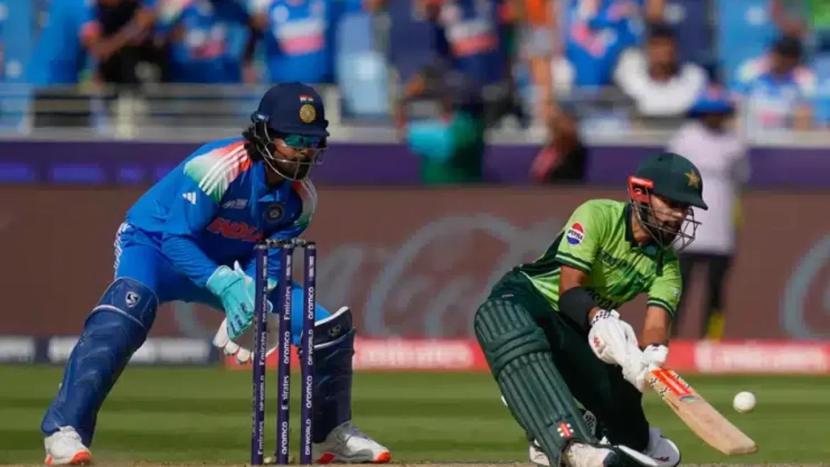 A cricket match scene showing an Indian wicketkeeper in blue gear poised near the stumps as a Pakistani player in green attempts to play a shot.