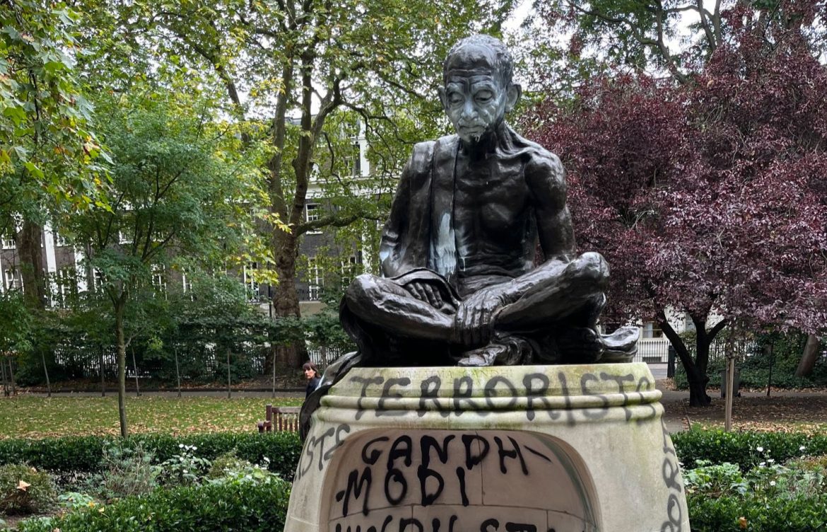 A bronze statue of a seated figure in a park, featuring graffiti with political messages and surrounded by greenery.