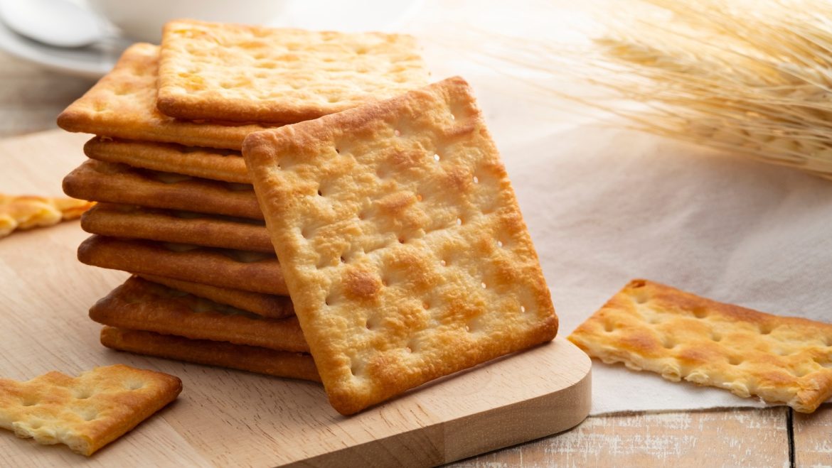 A stack of golden-brown crackers on a wooden cutting board, with a few scattered nearby, set against a soft, bright background.
