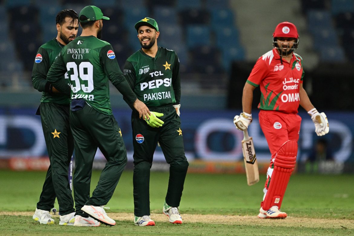 A group of Pakistani cricketers in green jerseys discuss strategy as an Omani player in red prepares to bat, set on a cricket field.
