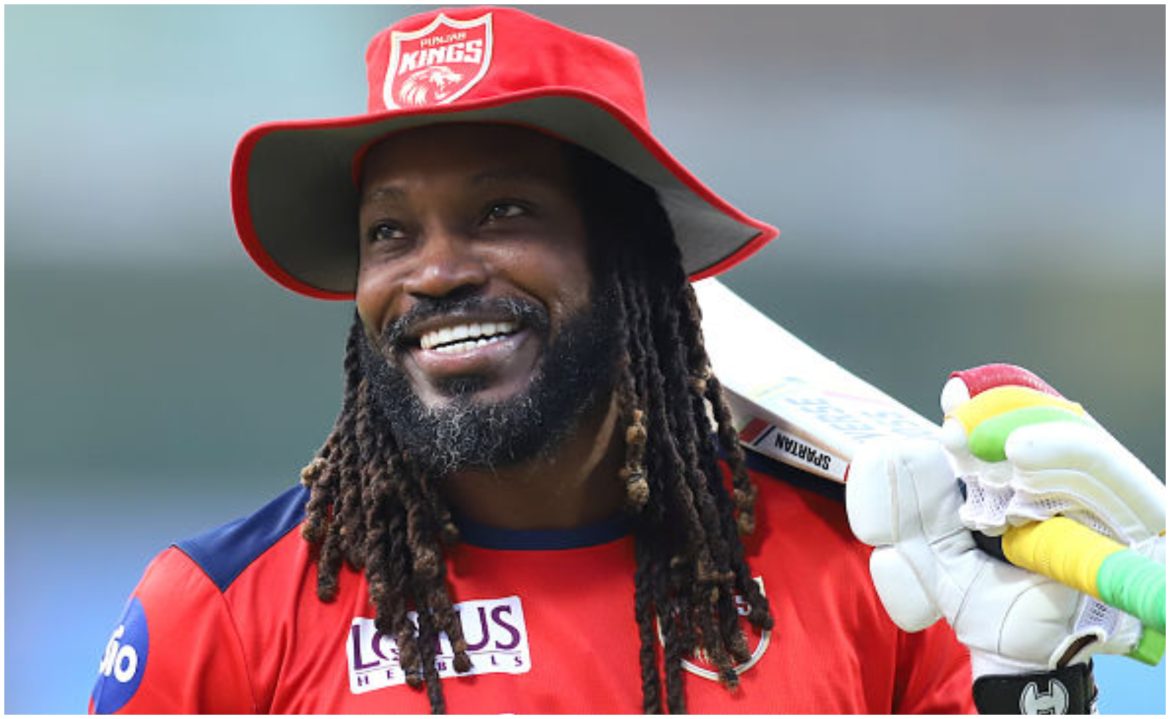 A cricketer in a red Punjab Kings jersey and a wide-brimmed hat holds a cricket bat, preparing for the game.