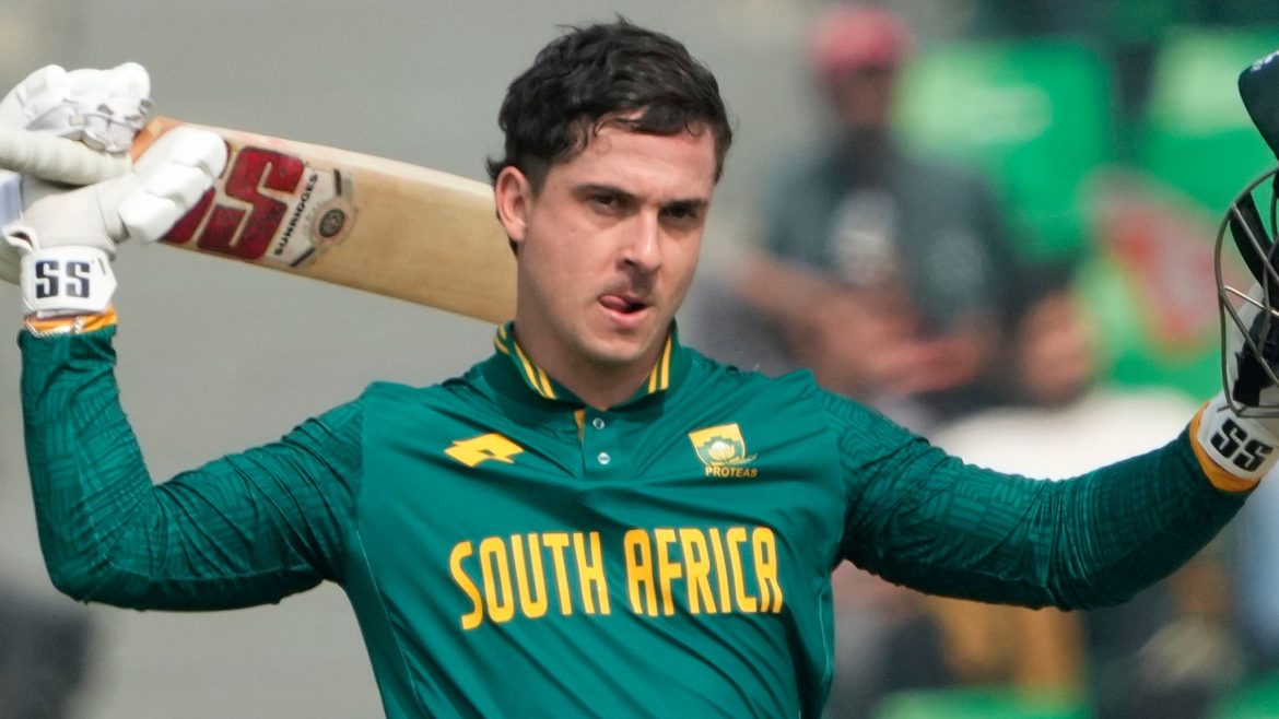 A South African cricketer in a green and gold uniform raises his bat in celebration, with blurred fans in the background.
