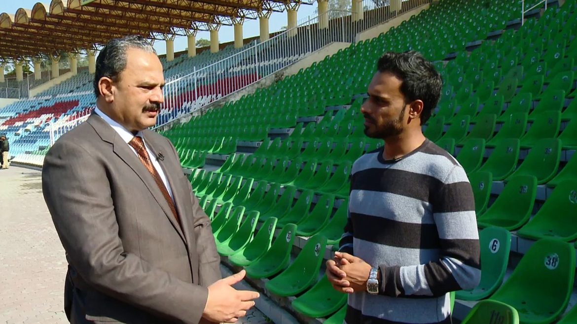 Two men engage in conversation at a nearly empty sports stadium, surrounded by green seats under a bright blue sky.
