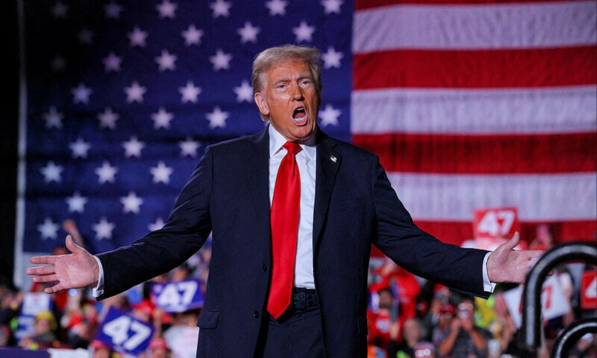 A man in a suit and red tie gestures to the crowd, standing in front of a large American flag at a political rally.