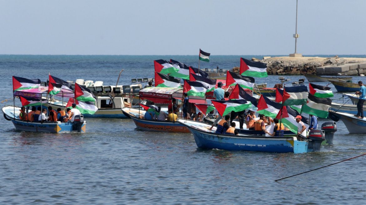 A vibrant scene of multiple boats on the water, prominently displaying Palestinian flags, with people engaging in an outdoor gathering.