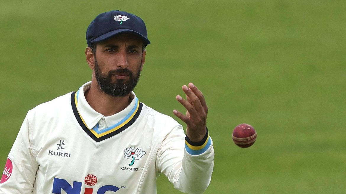 A cricketer in a white Yorkshire jersey prepares to catch a red cricket ball, focused and poised on the green field.