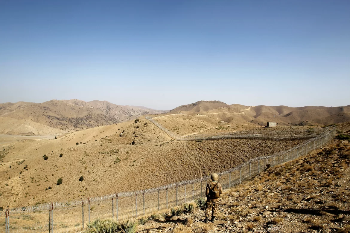 Pakistan Afghanistan Conflict - A soldier stands beside a barbed wire fence on a barren, mountainous border under a clear blue sky, conveying a sense of isolation and vigilance.