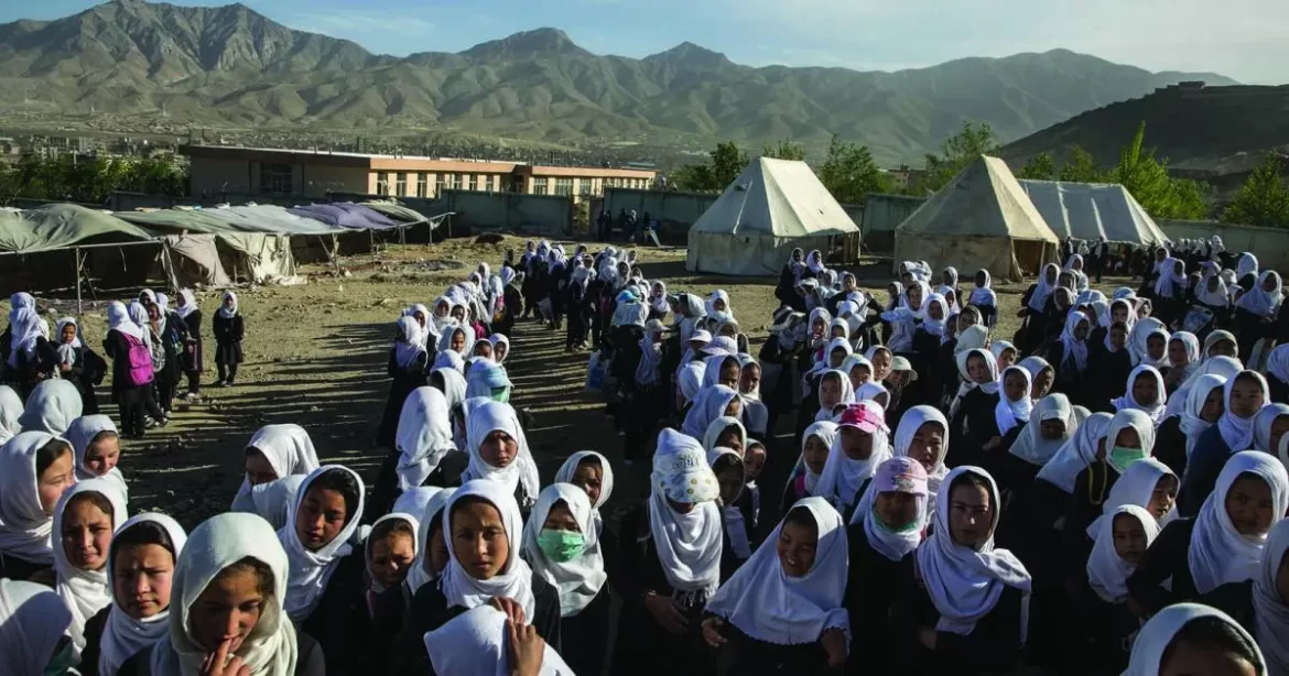 A large group of girls in traditional dress and headscarves assembles outdoors, with mountains and tents in the background.