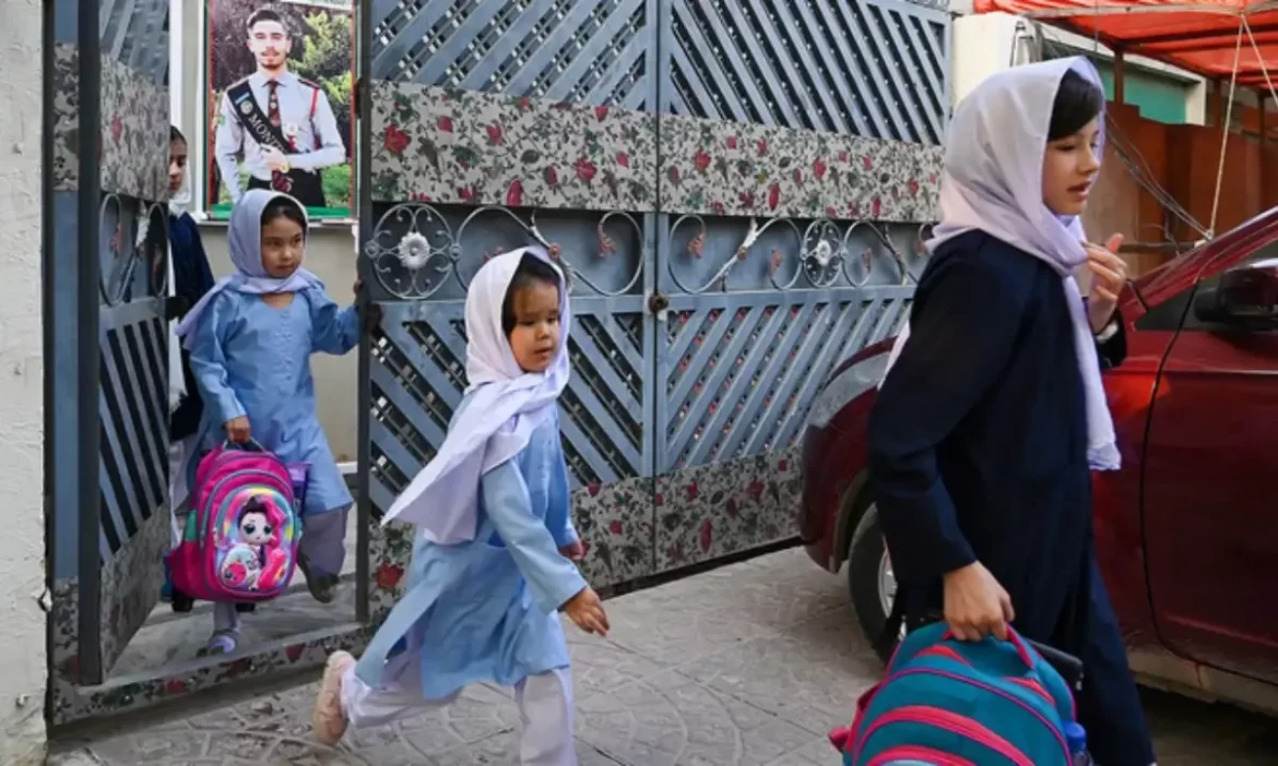 Young girls in school uniforms carrying colorful backpacks walk through a decorative gate, ready for school. A portrait hangs above the gate.