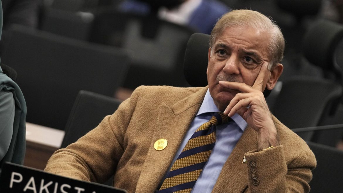 A well-dressed individual in a beige blazer and striped tie seated at a conference table, with a nameplate reading Pakistan.